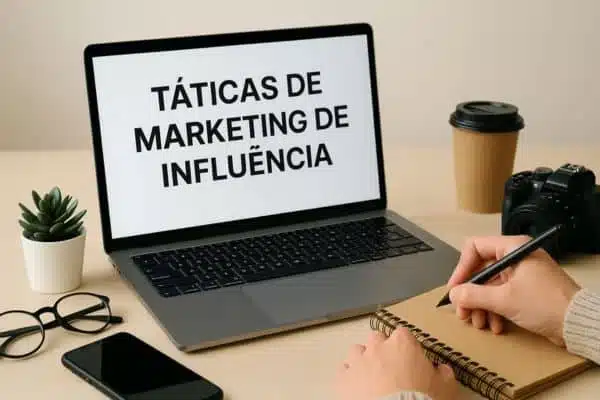 Um notebook em cima da mesa e nele esta escrito Táticas de Marketing de Influência na sua frente aparece as mãos de uma pessoa escrevendo com uma caneta em um caderno