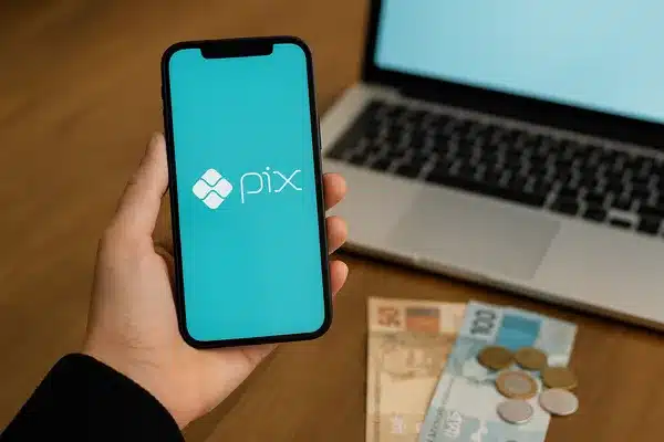 Ganhar dinheiro no Pix em 2025Pessoa segurando smartphone com tela do Pix, dinheiro em notas e moedas ao lado de um notebook.