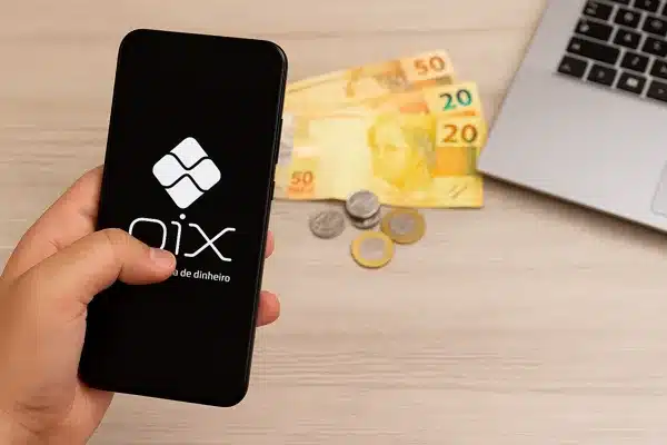 Ganhar dinheiro no Pix em 2025Mão segurando smartphone com logo do Pix, notas e moedas brasileiras ao lado de um notebook.
