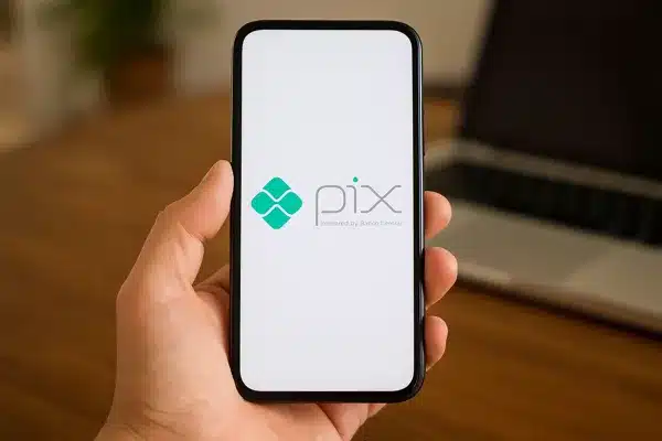 Ganhar-Dinheiro-No-PixPessoa segurando um smartphone que exibe um logotipo verde na tela, com um notebook desfocado ao fundo sobre uma mesa de madeira.