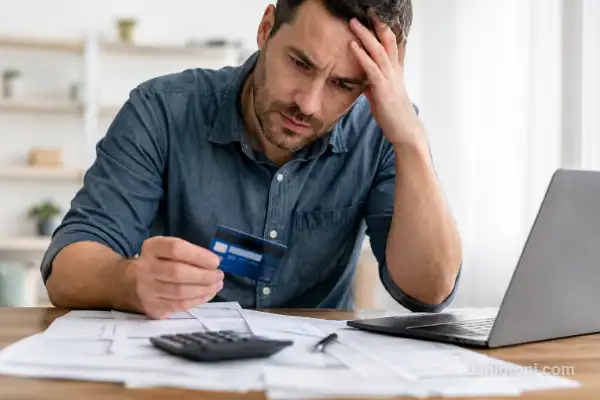 atrasar o cartão em 2026 com homem preocupado segurando cartão de crédito diante de contas, calculadora e notebook
