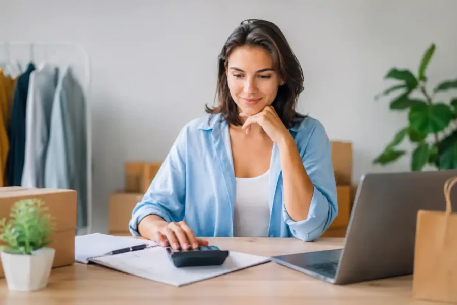 Empreendedora usando calculadora ao lado de notebook e caixas de produtos para calcular o capital de giro para MEI.